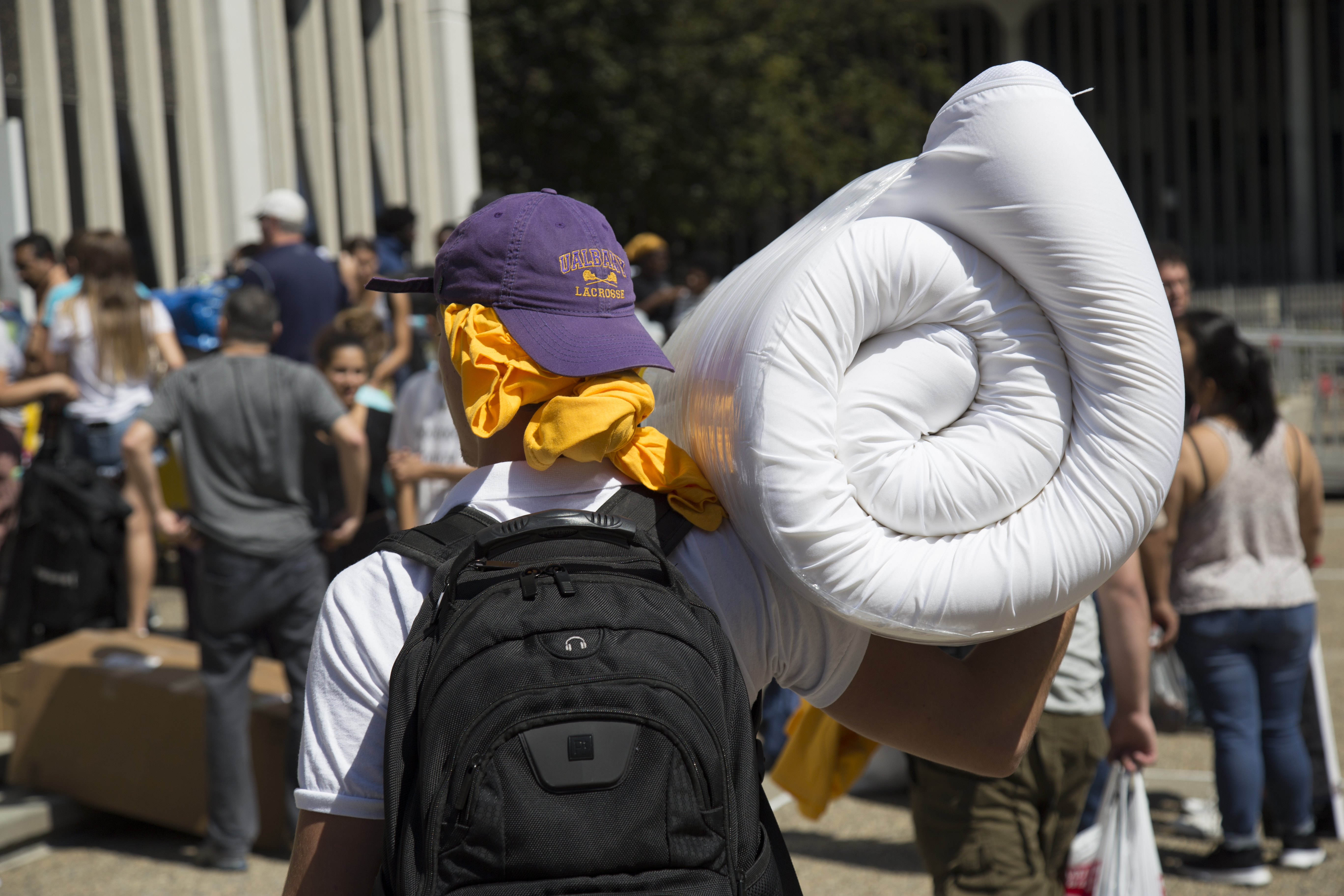 A student holds a mattress topper on their shoulder, wearing a UAlbany lacrosse hat and black backpack