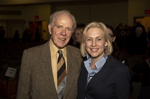 An older man in a tan suit coat and yellow shirt with a striped tied poses for a photo with a blond woman in a blue collared shirt and dark coat.