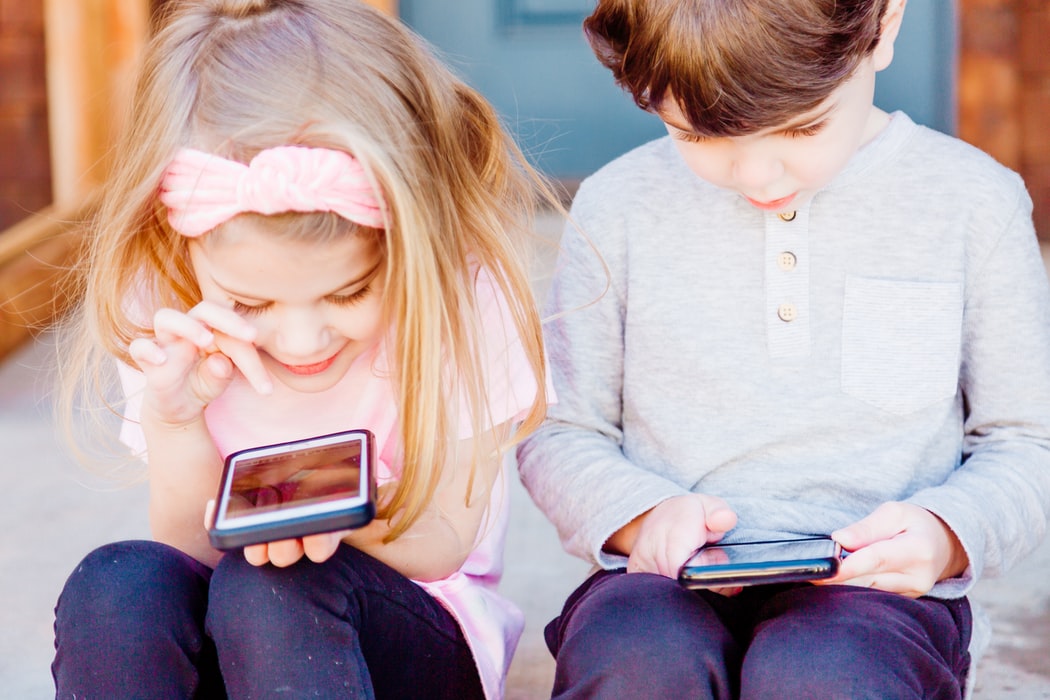 A blond girl in a pink shirt and jeans and a boy in a grey shirt and jeans sit outside on a step, playing on cell phones.