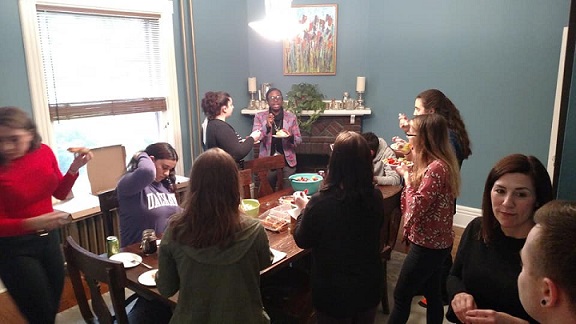 Eleven people sit and stand around a dining room table, smiling, talking and enjoying food.