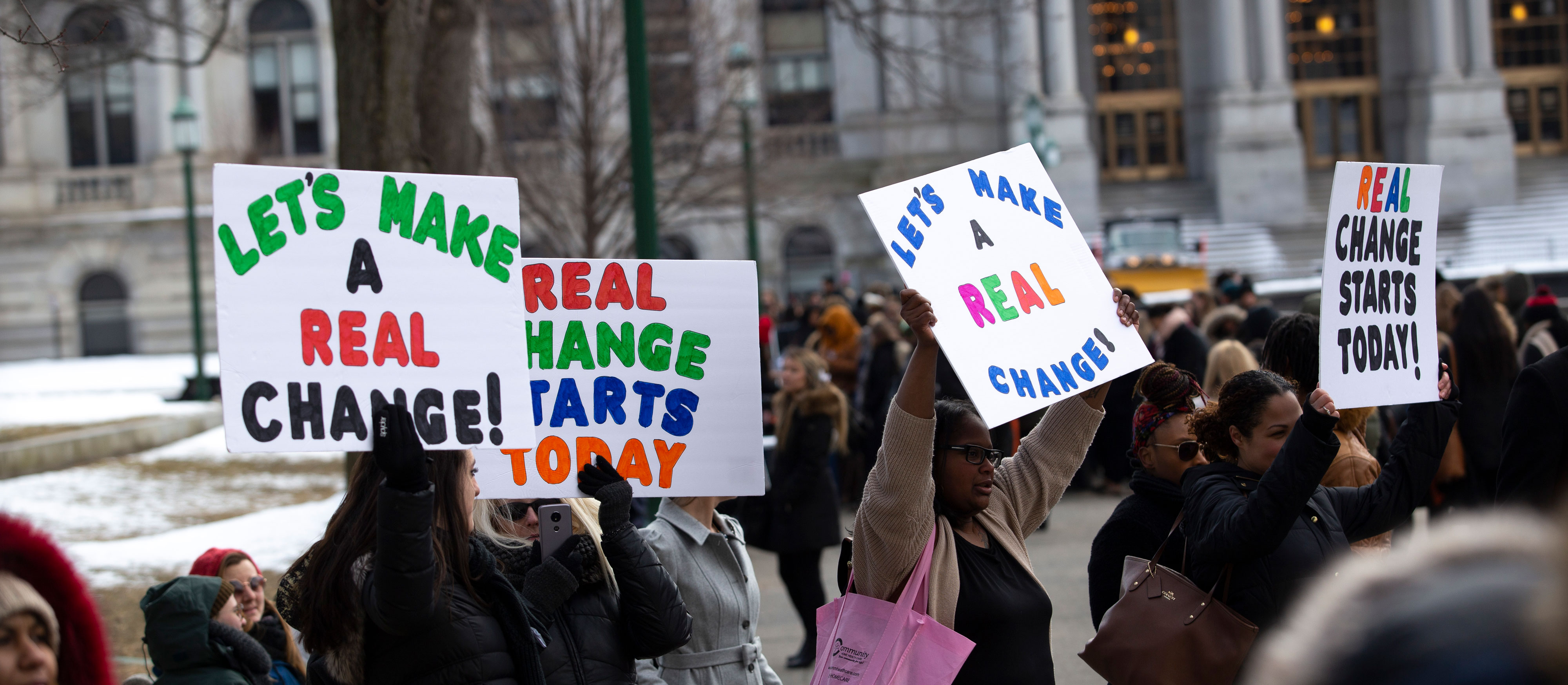 Students and instructors hold up "Let's make real change!" and "Real Change Starts Today" signs at a protest outside the New York State Capitol.