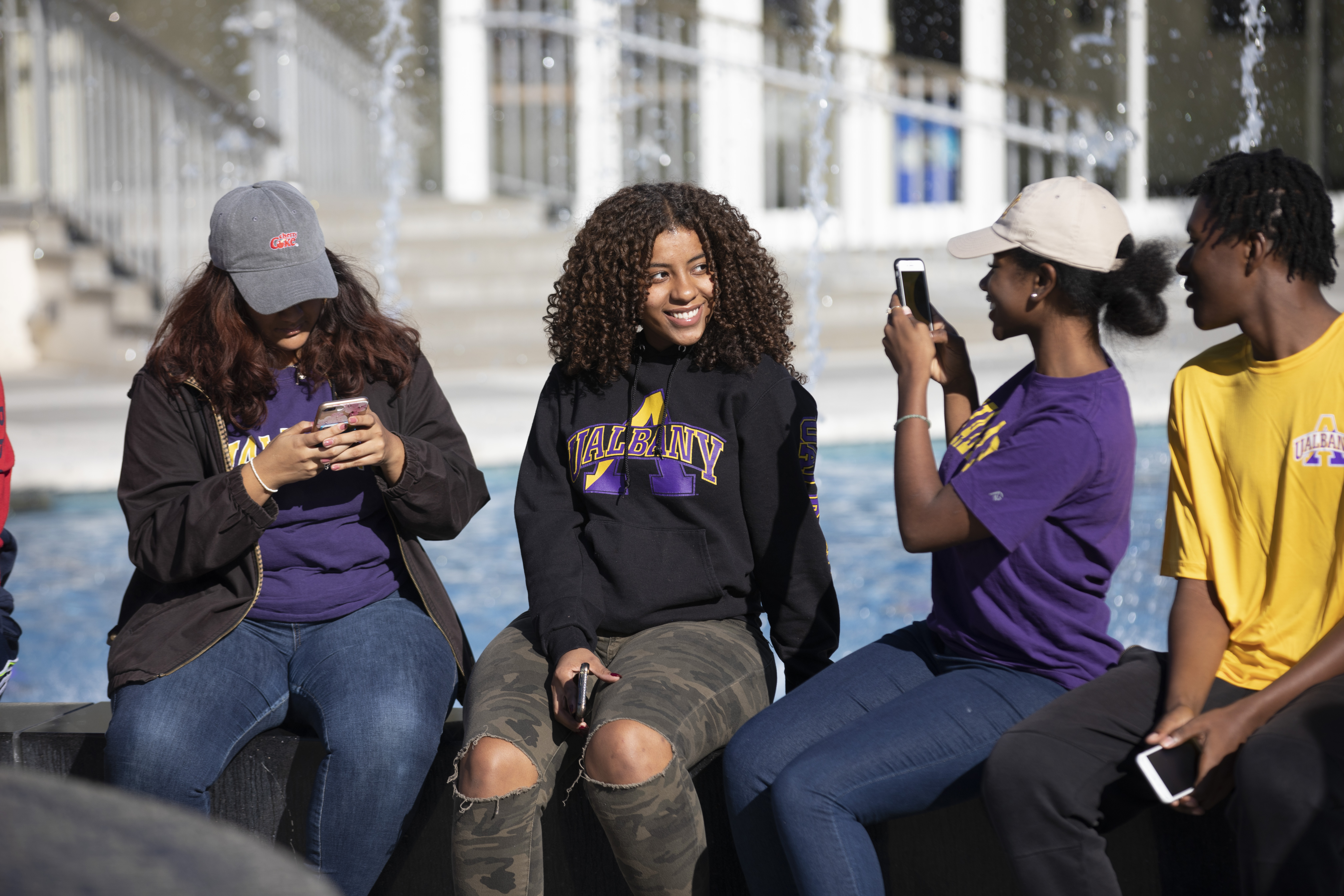 Four students sit on the edge of a fountain, one poses for a photo that the other is taking