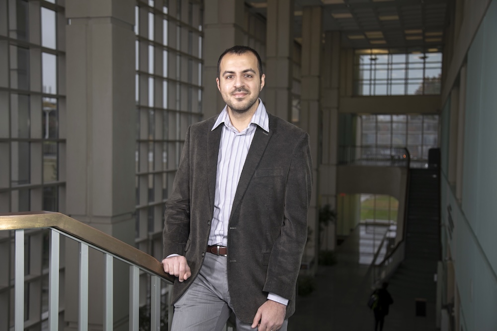 A man in a blazer leans against a railing as he poses for a portrait in a glass-walled building.
