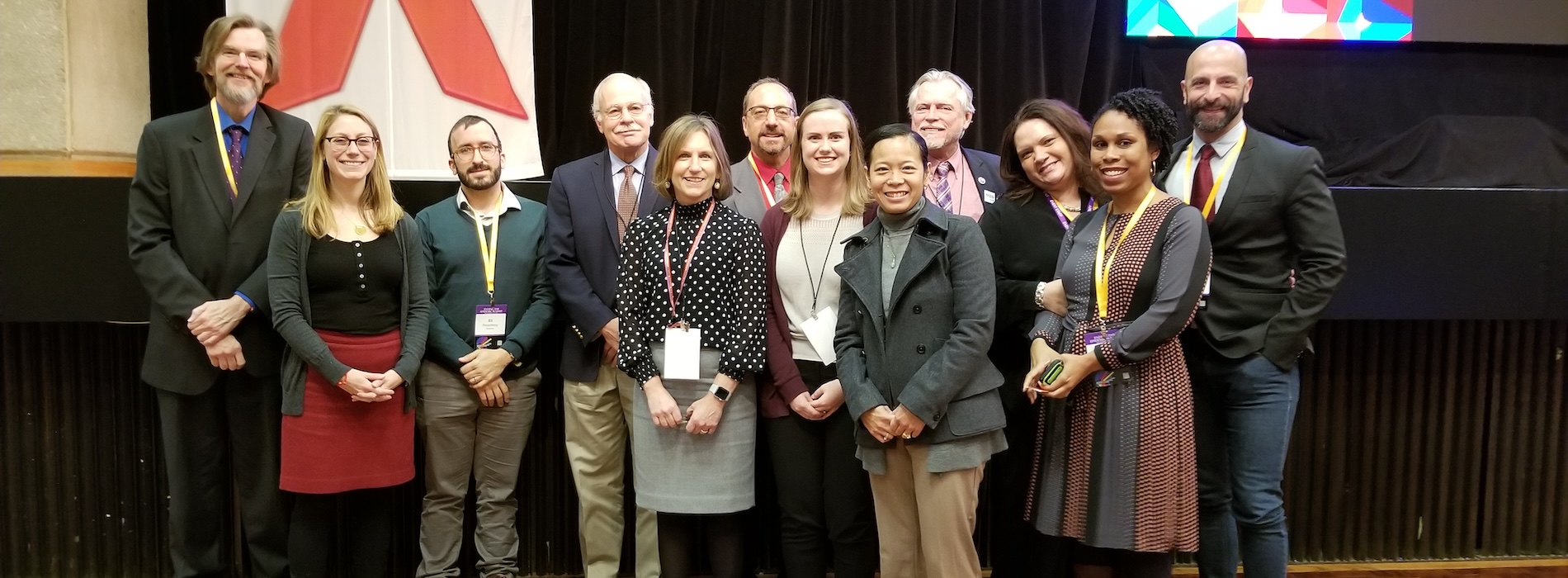 A group of CCHRPP researchers stand in front of a black wall. A white flag hangs on the wall behind them. On the flag is the red HIV/AIDS ribbon.