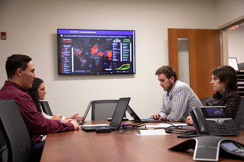 Researchers gather around a table with laptops, next to a large screen showing a map, as they work together in the VOST lab.