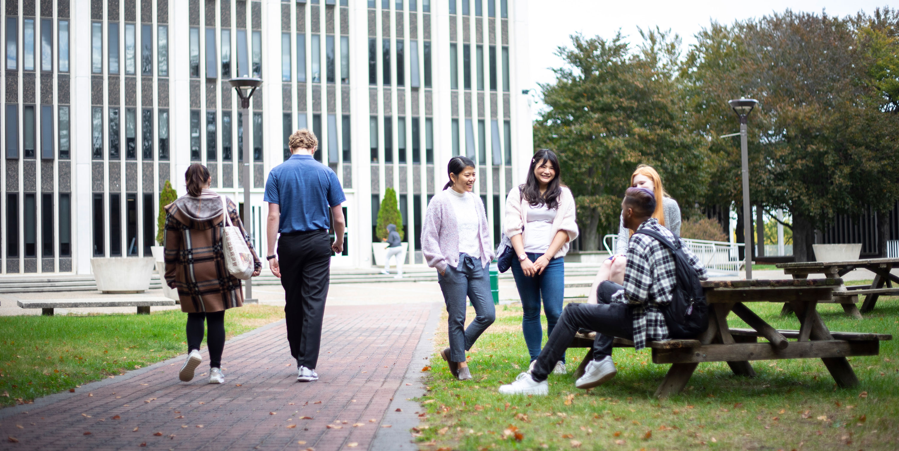 Two students walk along a campus pathway, passing a group of four other students gathered around a picnic table talking.