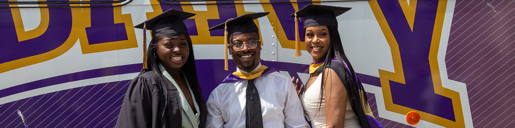 Three graduating students standing in front of a UAlbany bus.