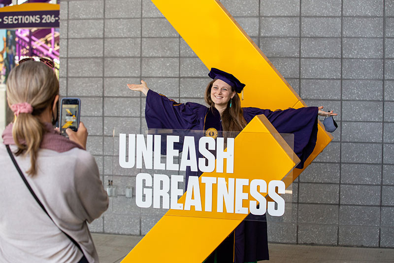 Student in Regalia Standing in the Unleash Greatness Chevron
