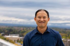 A smiling portrait of a man wearing a blue collared shirt on a rooftop against a blurred background of trees and mountains.