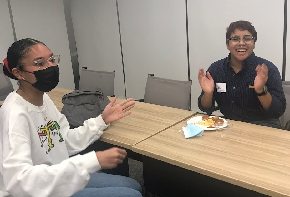 Two students at a conference table wearing glasses and clapping, one in a mask and white sweatshirt, the other in a black shirt and smiling