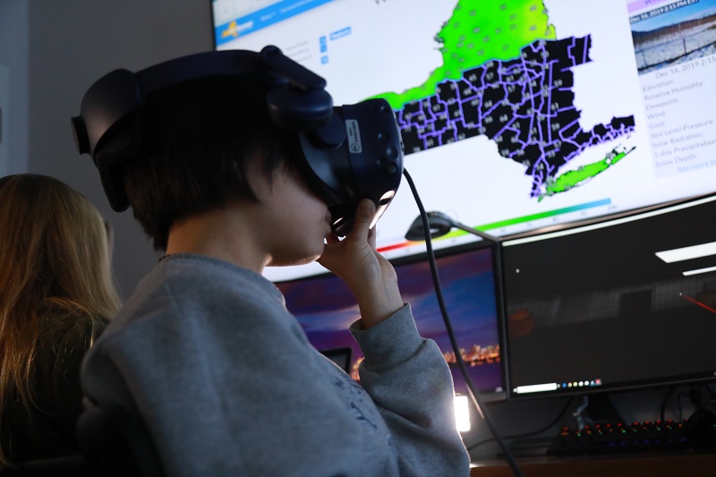 A profile photo of a student wearing VR googles in the foreground and backlit by a large wall-mounted screen displaying a New York State Mesonet weather map.