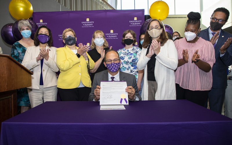 A masked President Rodriguez is seated at a purple table holding up a certificate while a crowd of masked people stand behind him and applaud.