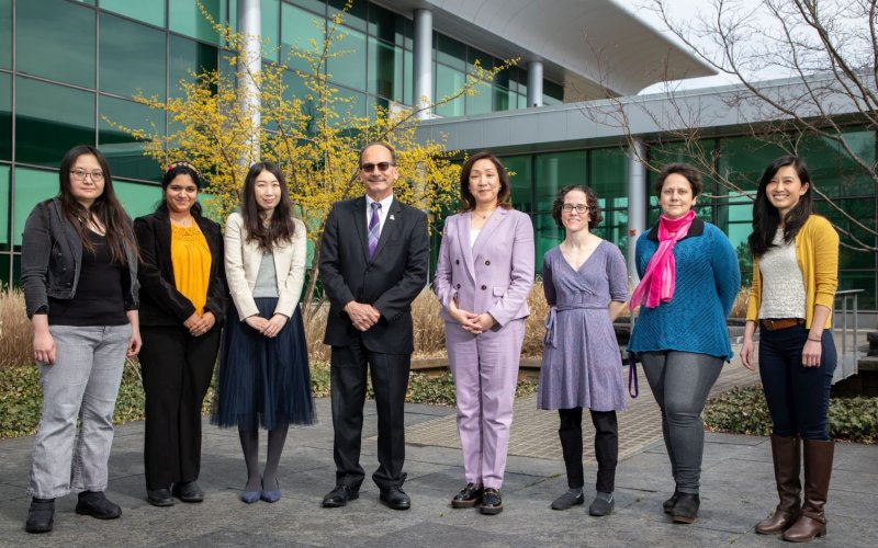 President Rodríguez and Provost Kim pose in a line with six Project SAGES researchers in front of greenery in the courtyard of the Life Sciences Research Building.