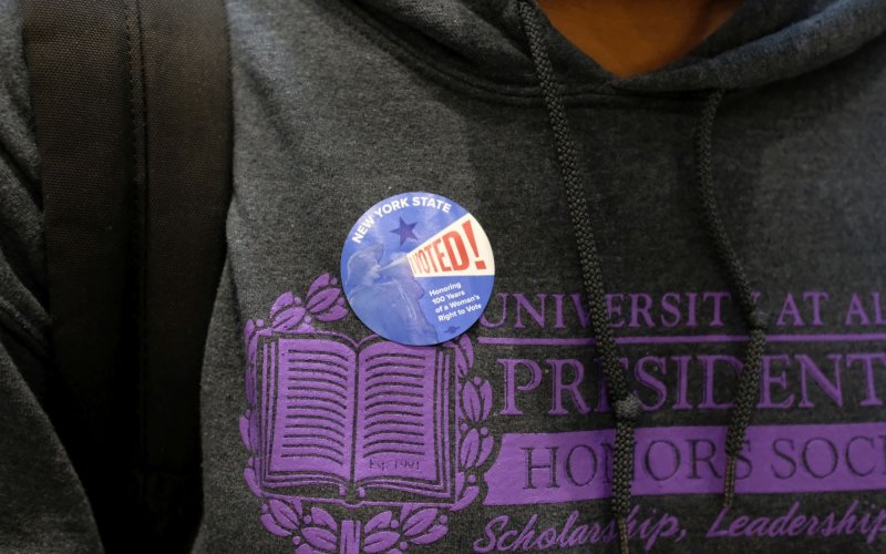 A close-up of an "I voted" sticker on a gray University at Albany shirt.