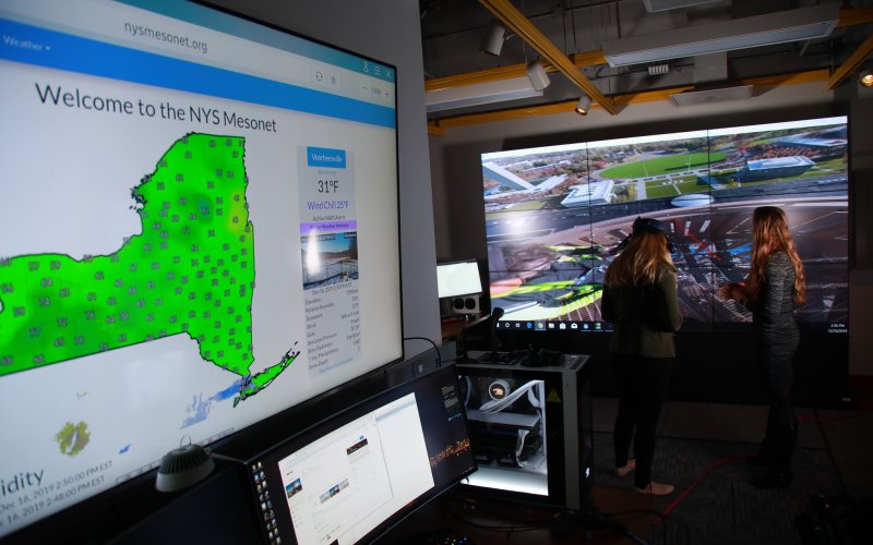 A photo showing screens in UAlbany's xCITE visualization lab, including one displaying a green map of New York and weather data from the New York State Mesonet.  