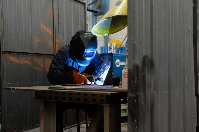 Sparks fly as a welder shrouded in protective gear, including a full mask, welds at a table in the Boor Sculpture Studio.