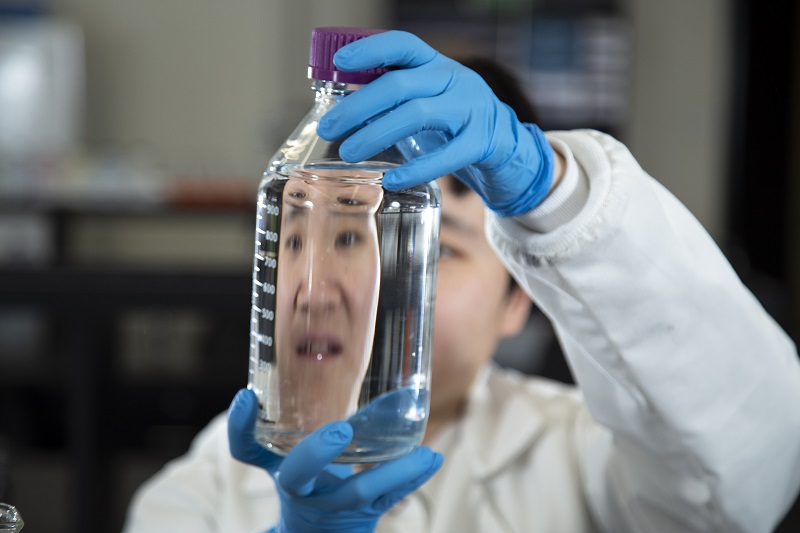 A student dressed in white lab coat and blue gloves peers through a glass laboratory bottle containing a clear liquid.