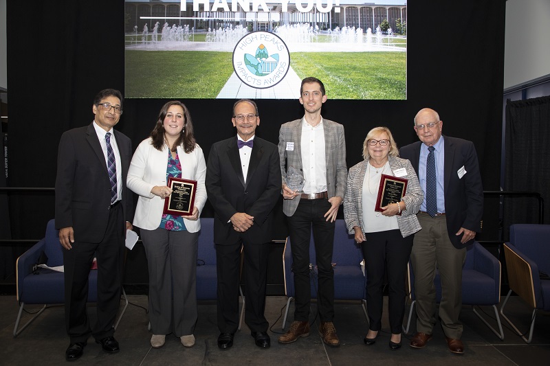 President Rodríguez and Dean Sen from the School of Business pose standing in a line with four representatives of the finalists for the inaugural High Peaks Impact Award. On the screen behind them, the HIPA logo is projected over an image of UAlbany's Entry Plaza fountain.
