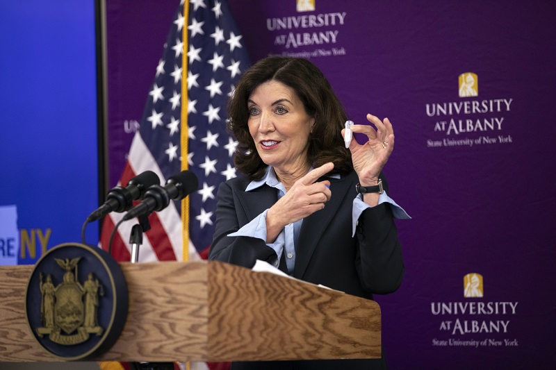 New York Governor Kathy Hochul stands at a brown podium bearing the seal of the State of New York holding a saliva tube from one of the RNA Institute's COVID-19 test kids.