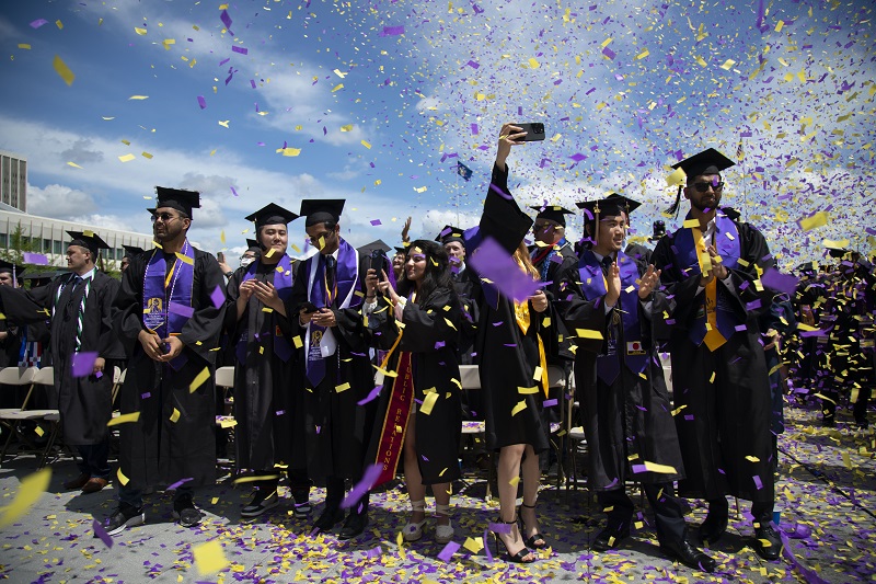 A crowd shot of students in graduation robes smiling and taking selfies amid a cloud of purple and gold confetti.