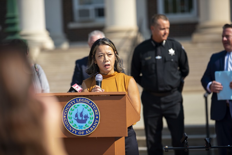 Associate Professor Tomoko Udo stands holding a microphone at a podium bearing the Albany County seal with several Albany County officials, including Sheriff Craig Apple, standing behind her as she speaks. 