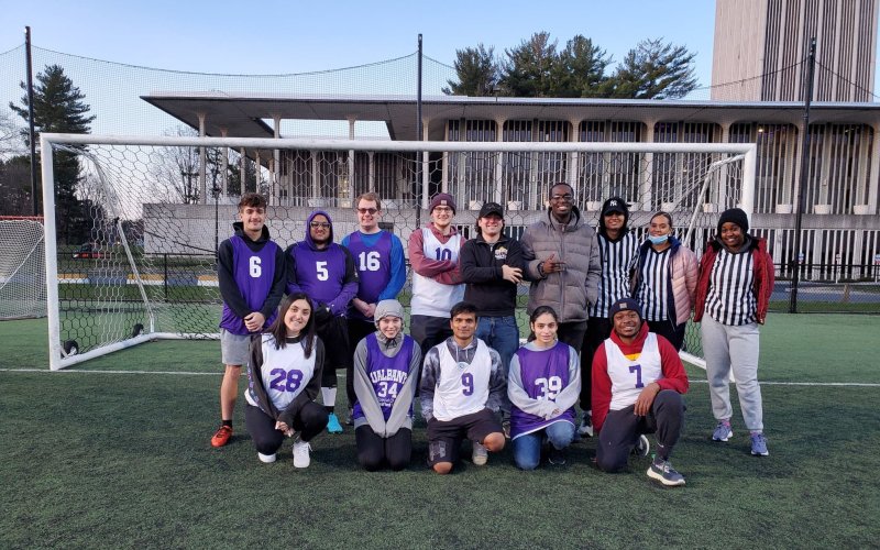 14 members of the UAlbany unified soccer league pose smiling in two rows in front of a soccer goal on the Dutch turf field with Dutch Quad in the background. nights this semester. (