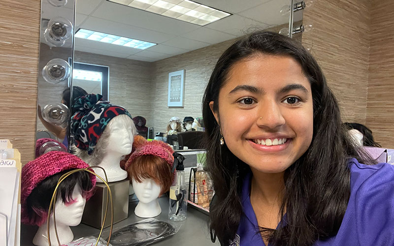 2022 Massry Fellow Labika Baral takes a selfie in front of a mirror and a display of wigs for patients at the American Cancer Society.