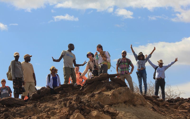 A group of younadults, some with hands in the air, stand on a rock outcropping in Kenya