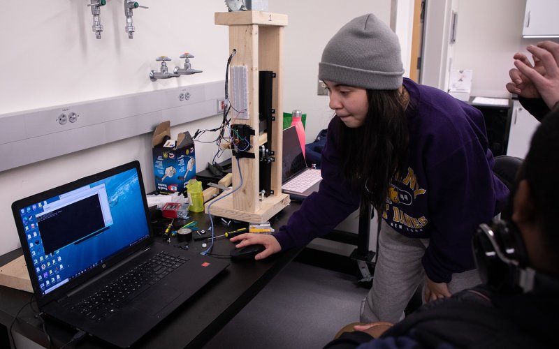 An engineering student in a gray knit cap and purple sweatshirt leans over a laptop on a workbench in an engineering classroom.