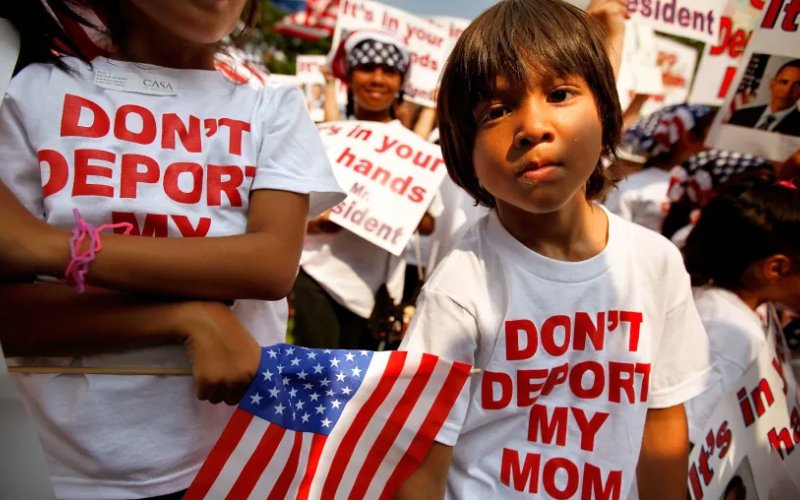 A young boy stands in front of a crowd of children, all wearing t-shirts that say "Don't Deport My Mom."
