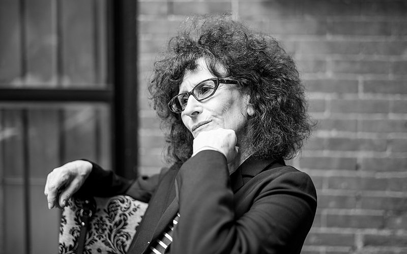 A black-and-white portrait of Lynne Tillman seated with her chin resting on her closed fist.