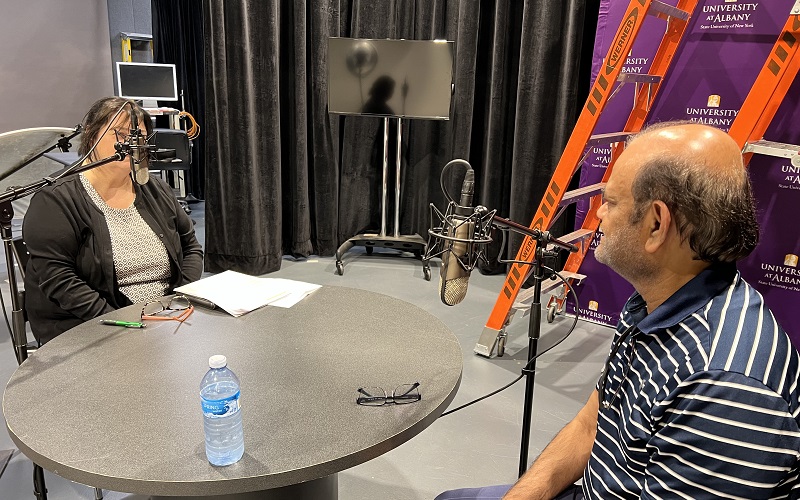 Podcast host Mary Hunt and VP for Research and Economic Development Kesh Kesavadas sit in front of microphones at a round table in the UAlbany digital media studio during a taping of The Engagement Ring. 
