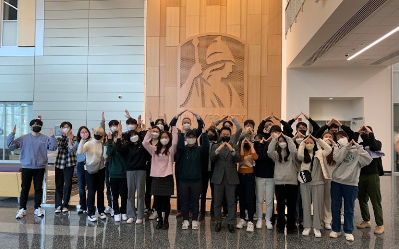 About two-dozen exchange students from South Korea pose with their arms in the air in the ETEC atrium in front of a wood-paneled wall depicting Minerva.
