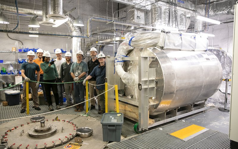 A group of scientists and movers stand near a large silver-colored cylinder dark matter tank inside of a lab.