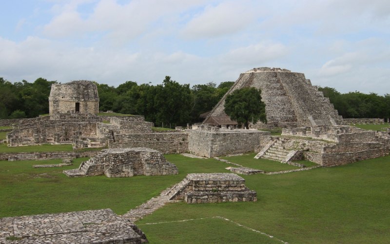 Square and pyramid shaped stone ruins of the ancient Mayapan city amid green grass.
