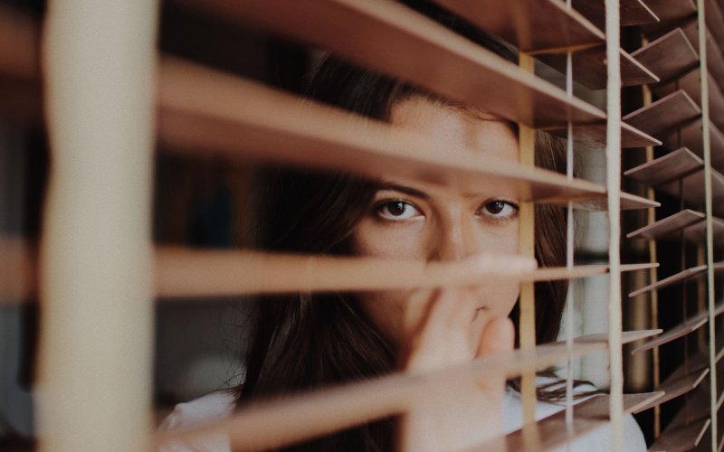 A woman peers through the wooden slats of window shades.