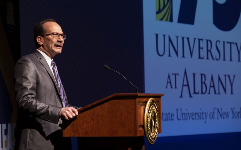 President Rodríguez, in a gray suit and purple striped tie, stands at a podium with the words University at Albany projected on a screen to his left. 