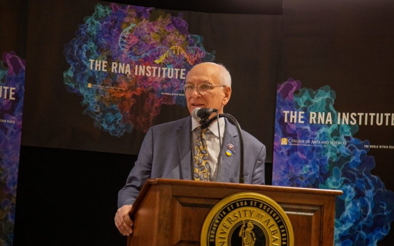 U.S. Rep. Paul Tonko stands facing the camera at a brown podium with a University at Albany seal addressing a crowd (not visible) in front of an RNA Institute banner.