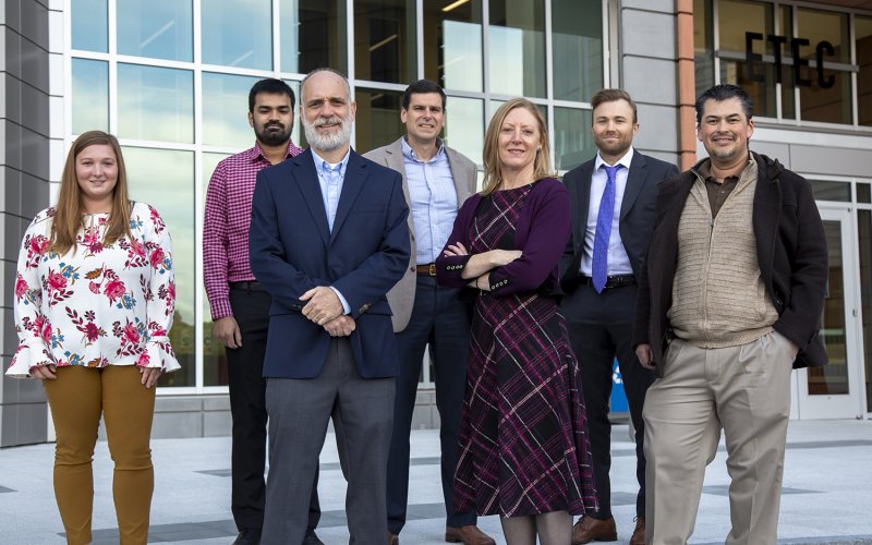 Seven members of the UAlbany research team stand in a group facing the camera in the plaza in front of the ETEC building.