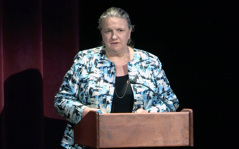 Dean Grimm stands at a podium with a red backdrop behind her wearing a blue and black jacket.