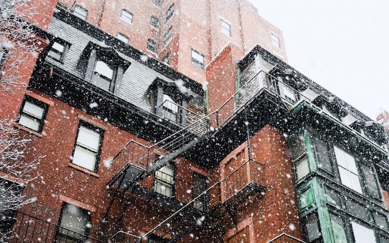 A brick apartment building with black fire escapes photographed from a low angle during a snow storm.