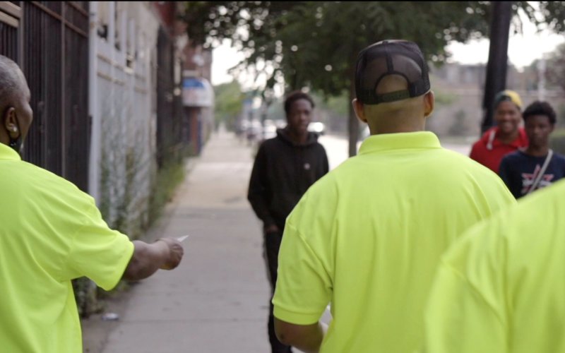 Men in yellow shirts, backs to camera, hand out pamphlets to some young men on the sidewalk