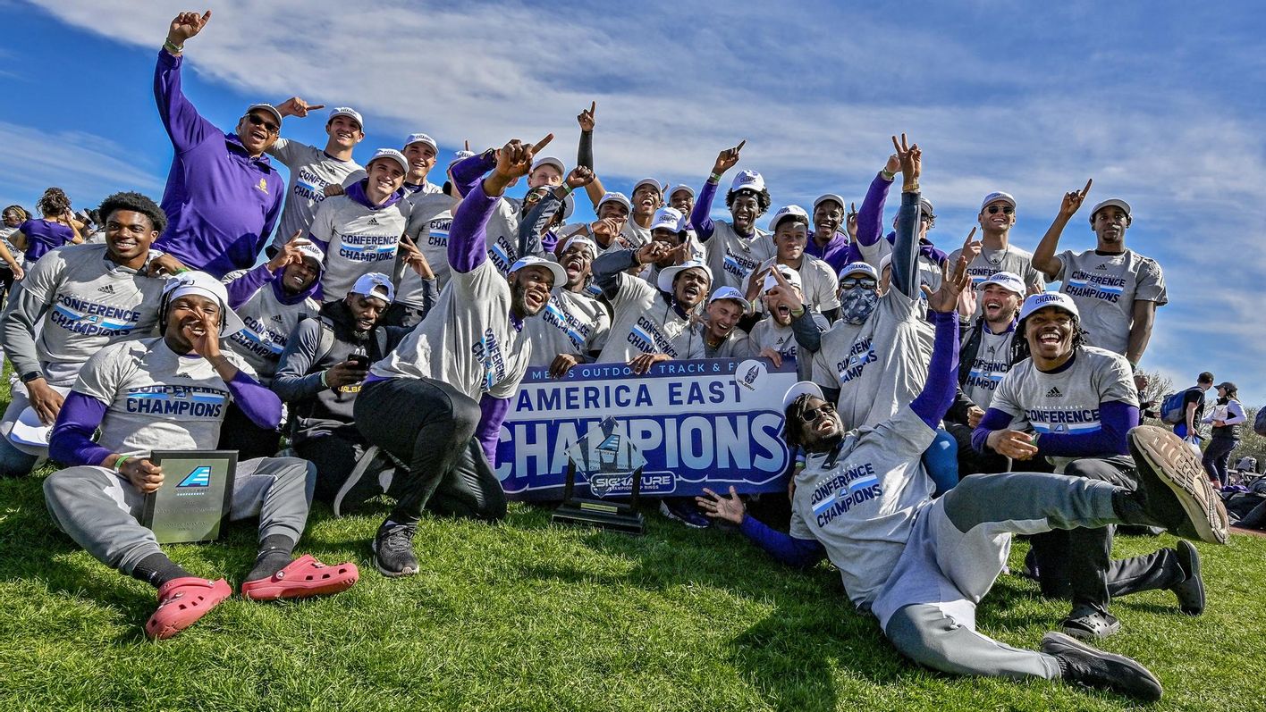 The men's track & field team, wearing gray "conference champions" t-shirts, sits in the grass with its arms and fingers in the air around a sign that says "America East Champions"