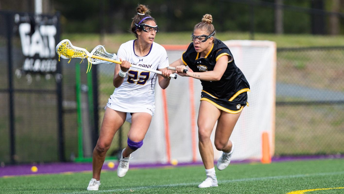 A UAlbany' women's lacrosse player wearing No. 23 and carrying the ball in her stick runs in mid-stride while being defended by a player from UMBC.