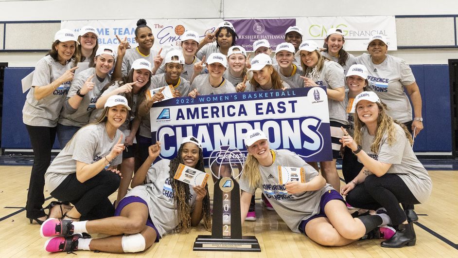 The Great Danes women's basketball team poses on the court next to the America East championship trophy holding a sign declaring them the 2022 America East champions.