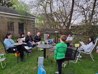 Seven people sit in chairs around a fire pit in a backyard, surrounded by trees and a house, with a child standing with his back facing the camera.