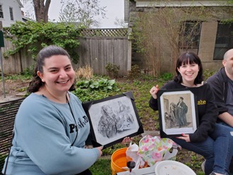 Two women with brown hair sit in a backyard, smiling while holding up framed historical drawings.