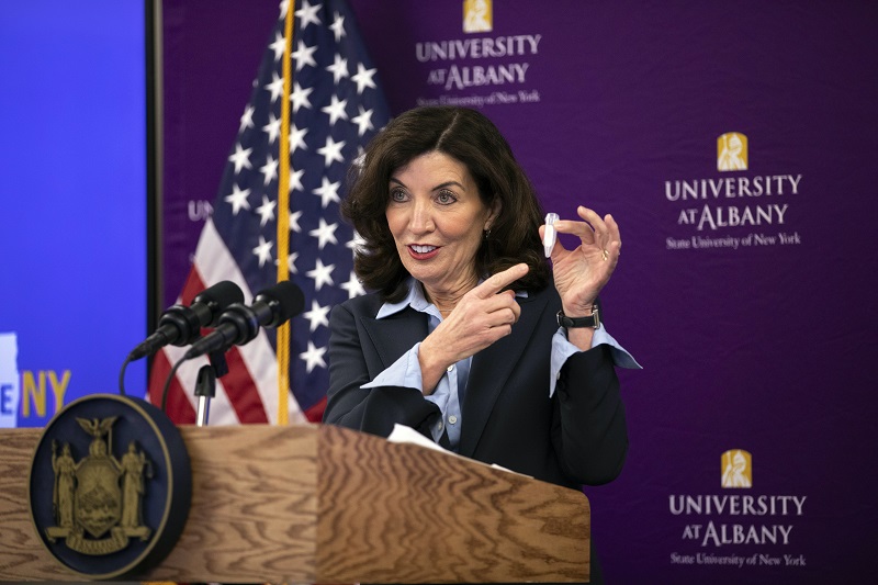 A woman standing at a lectern in front of a purple UAlbany banner holds up a vial used for saliva testing.
