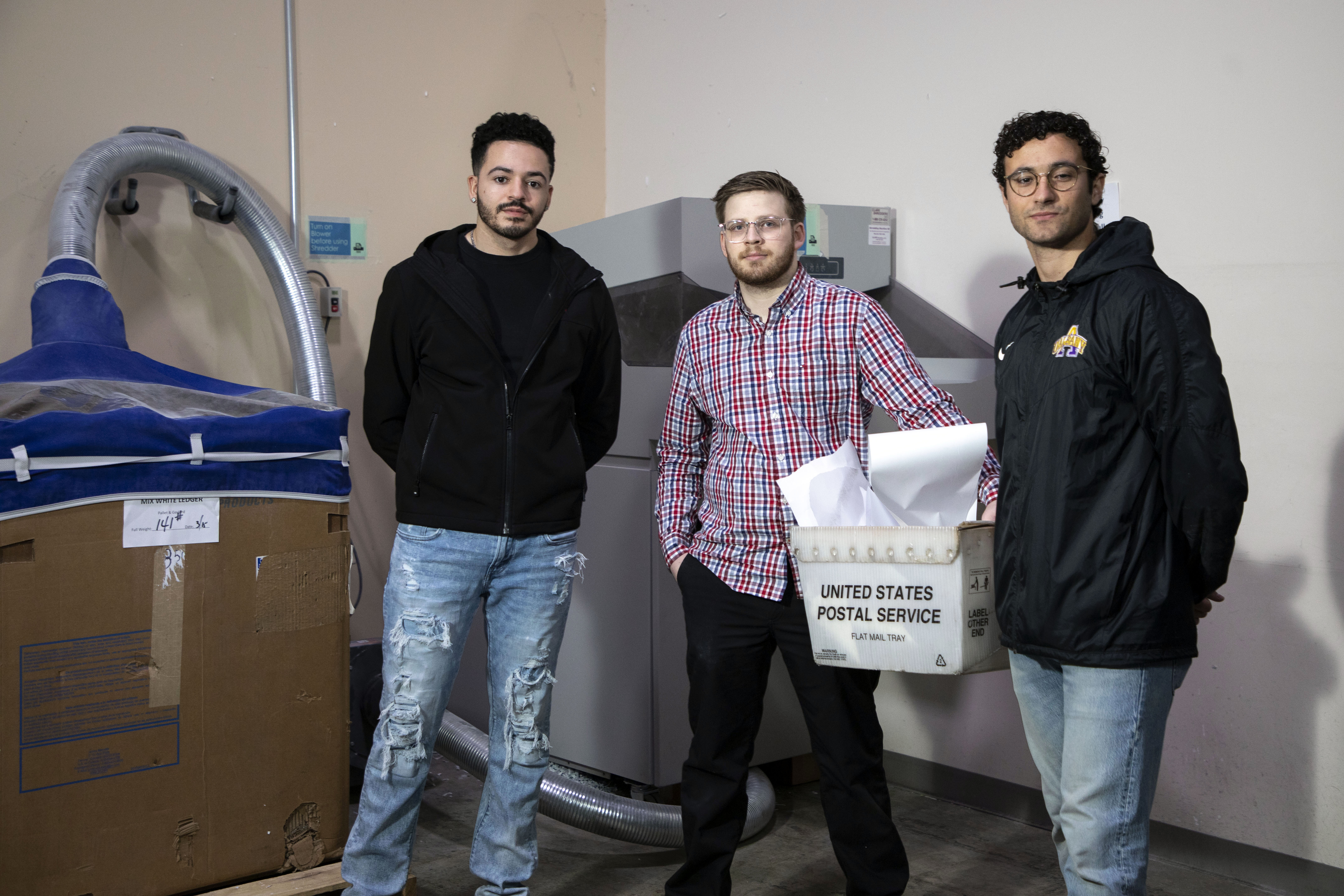 Three students stand indoors in front of a shredding machine and bypass that they created.