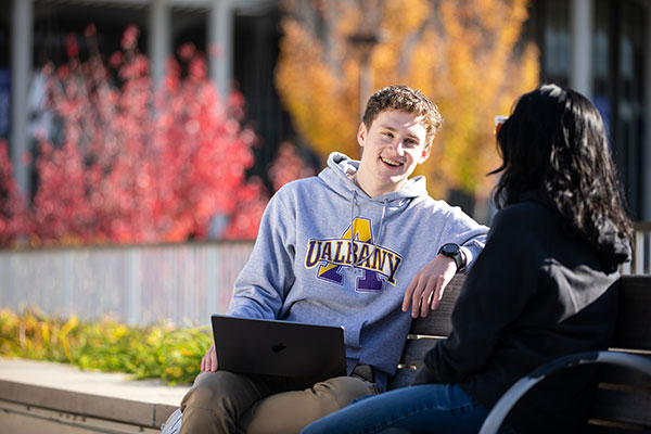 Two UAlbany students talk to each other while sitting outside on campus in the fall.